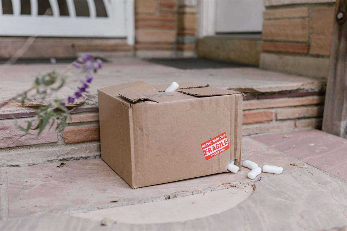 A dented cardboard box with a fragile sticker sits on porch steps with foam peanuts escaping