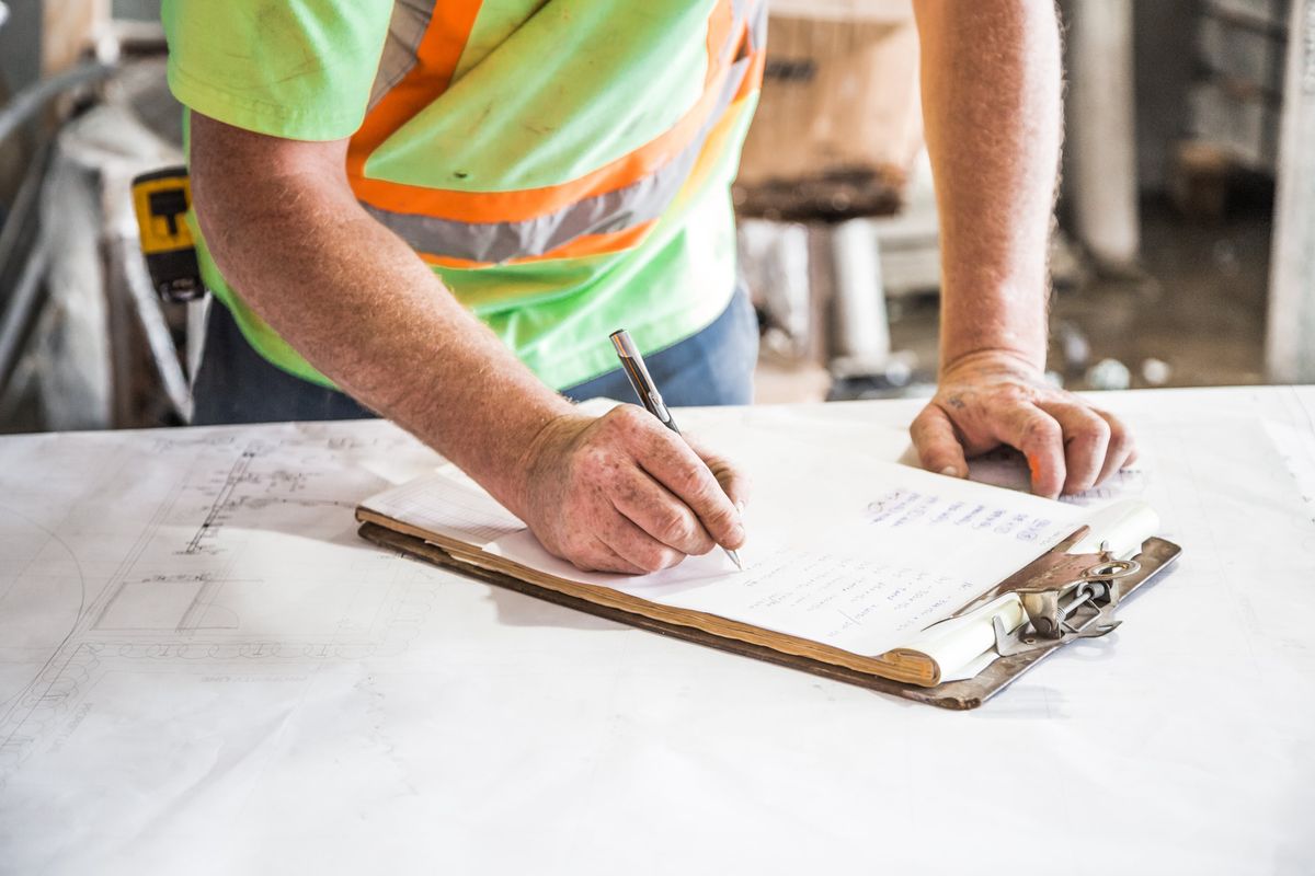 A builder in a high-visibility vest writes notes on a clipboard over site drawings