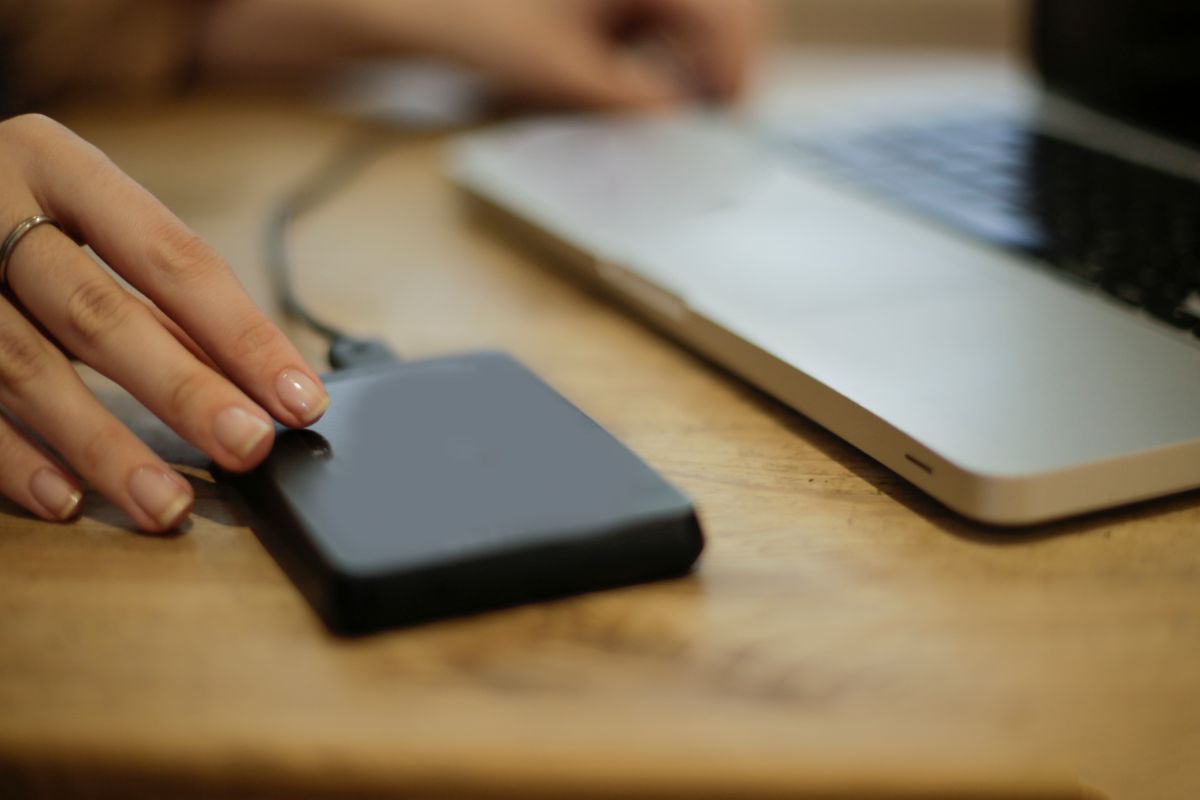 A hand resting on a small portable hard drive on a wooden desk beside an open laptop