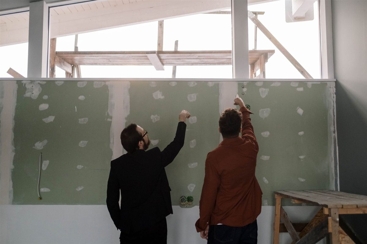Two people point at an unfinished plasterboard wall during a renovation discussion