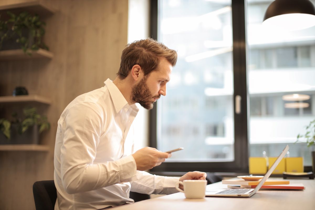 A person at an office desk checks their phone beside an open laptop and a cup of coffee