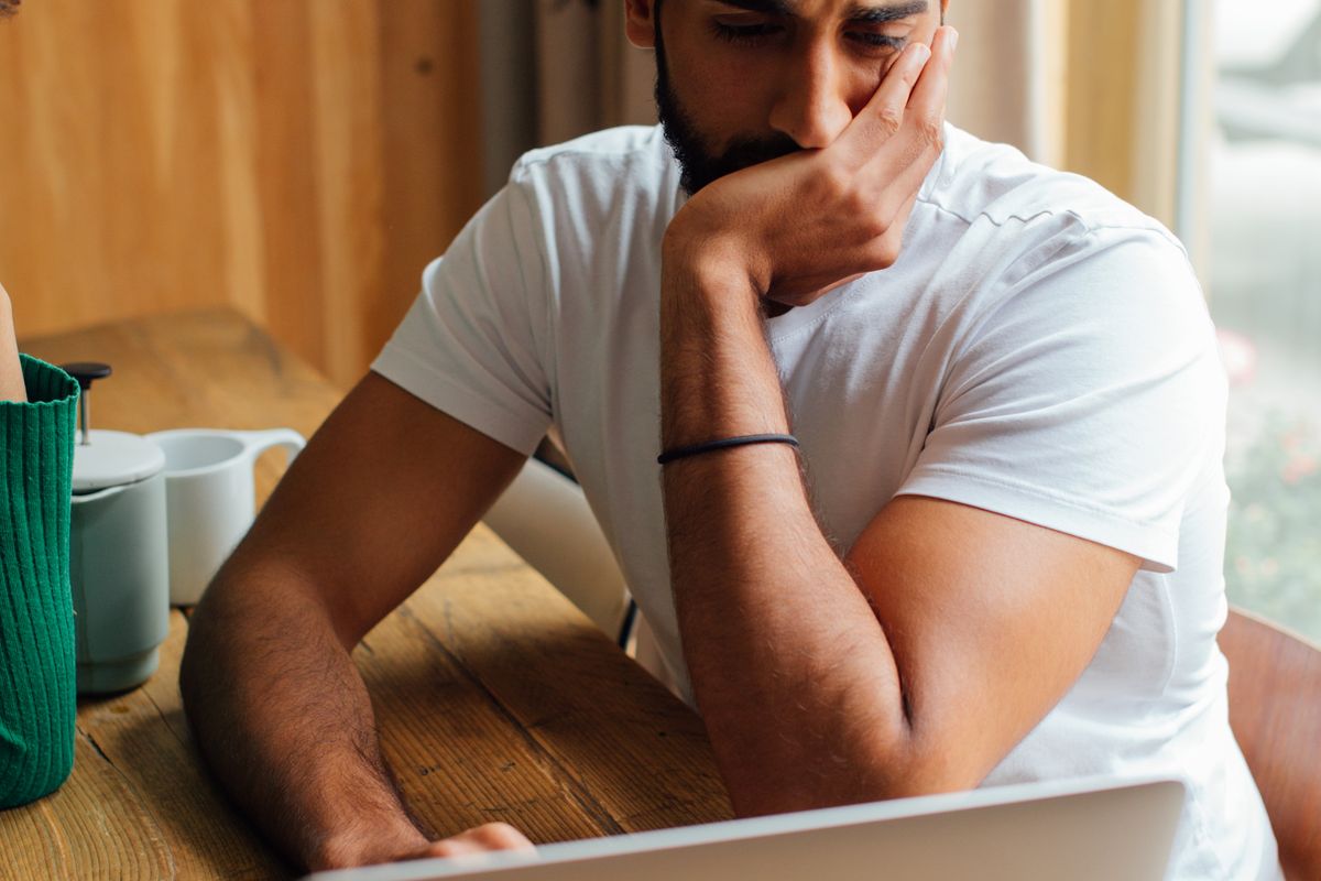 A person rests their chin on one hand while reading an unanswered email thread on a laptop at a wooden table