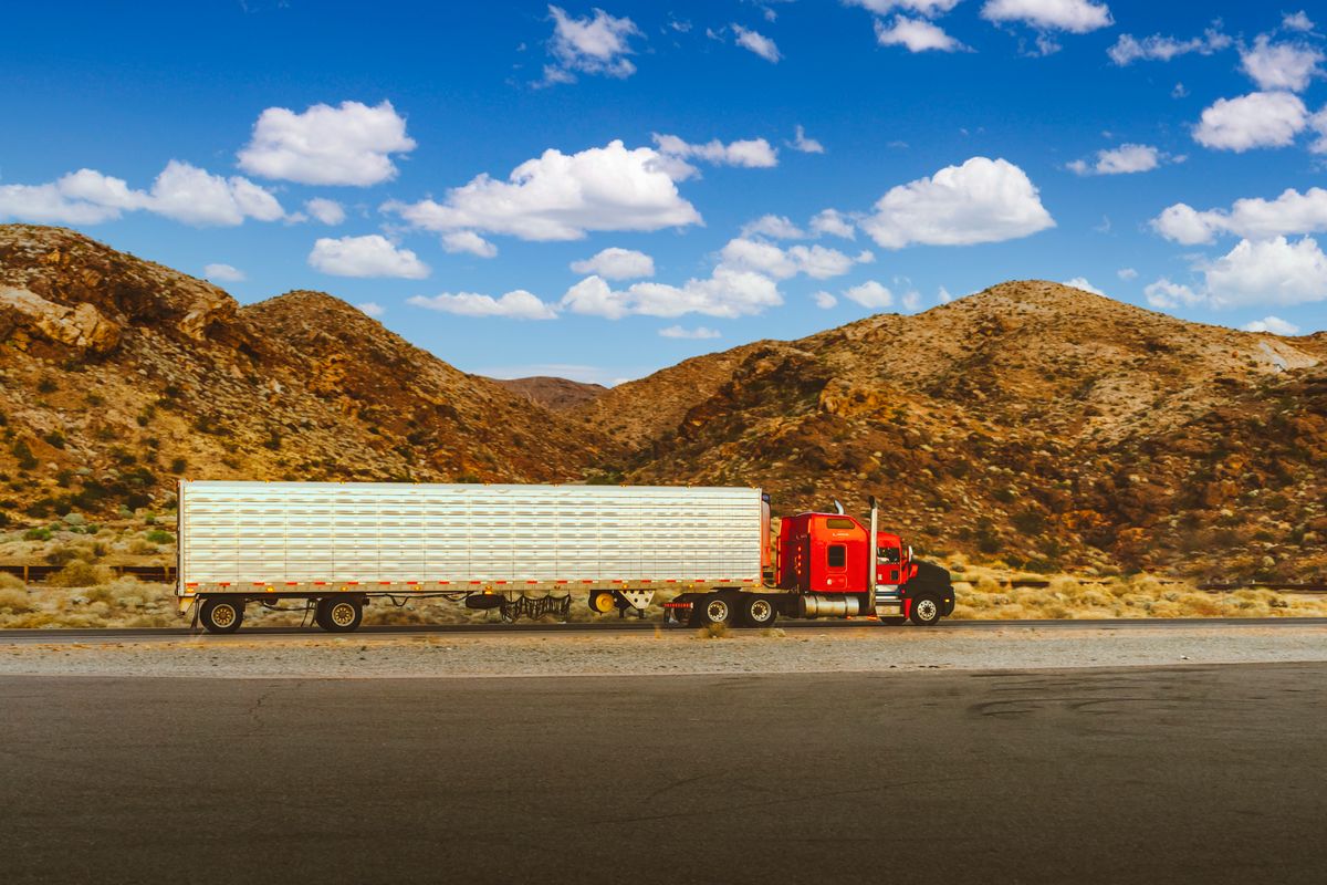 A red semi truck with a long trailer on a desert highway with mountains behind it