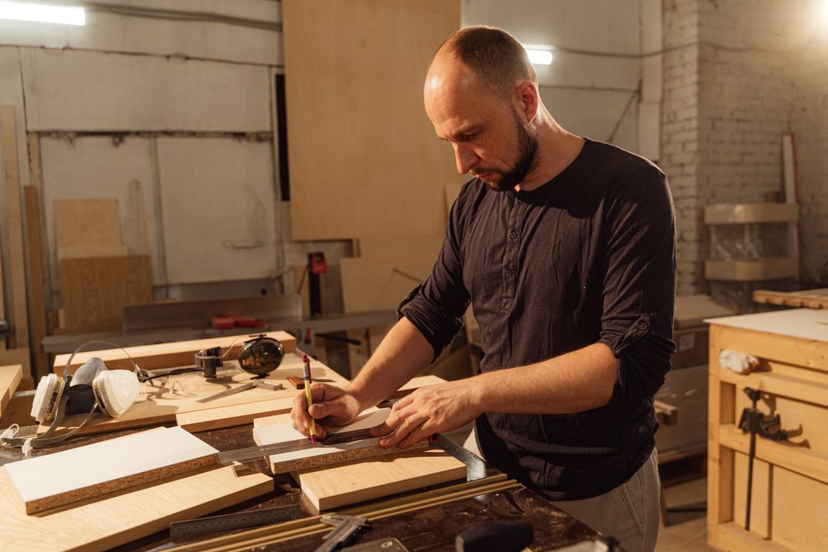 A carpenter measuring boards at a workshop bench, mid-job