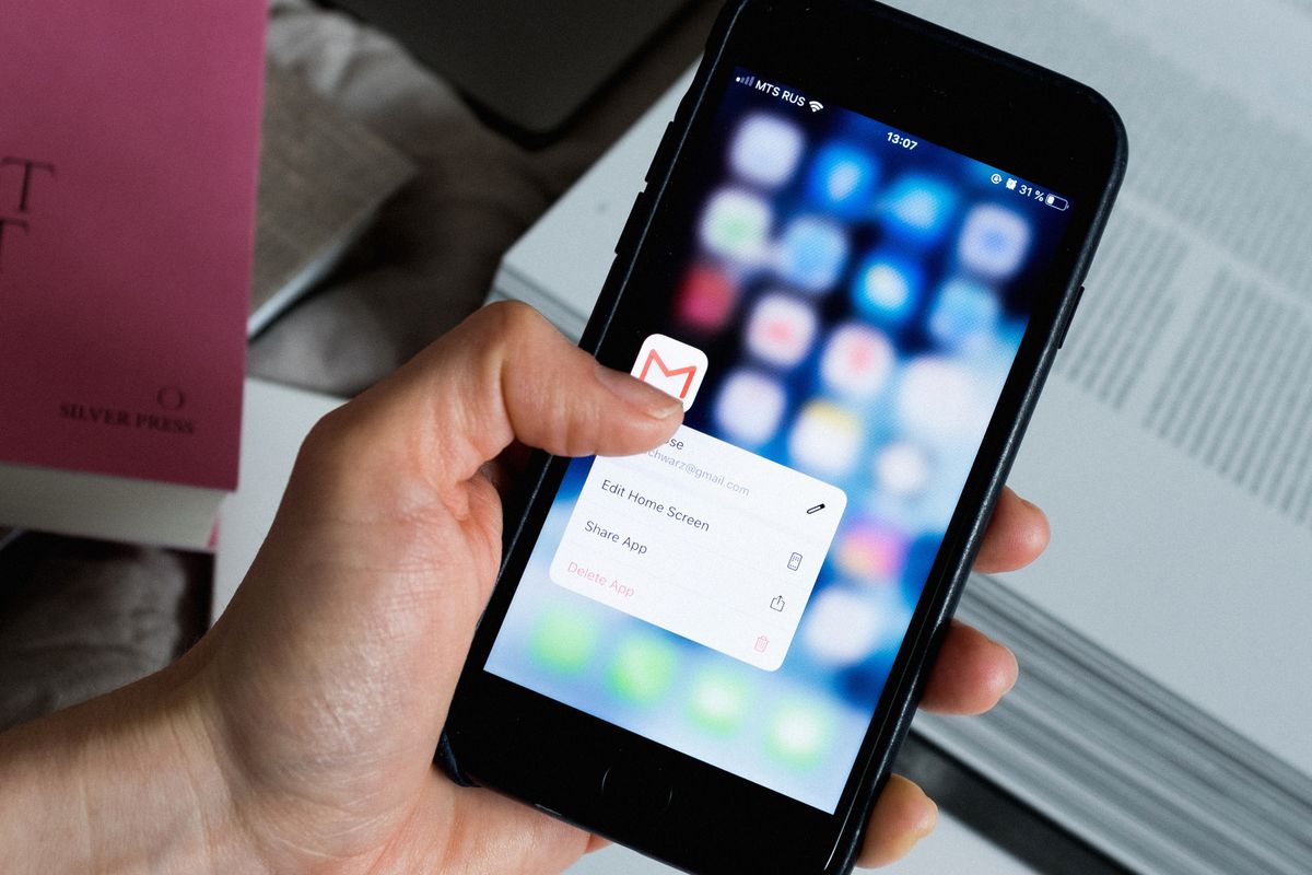 A hand holding a smartphone with a thumb pressed on the Gmail app icon