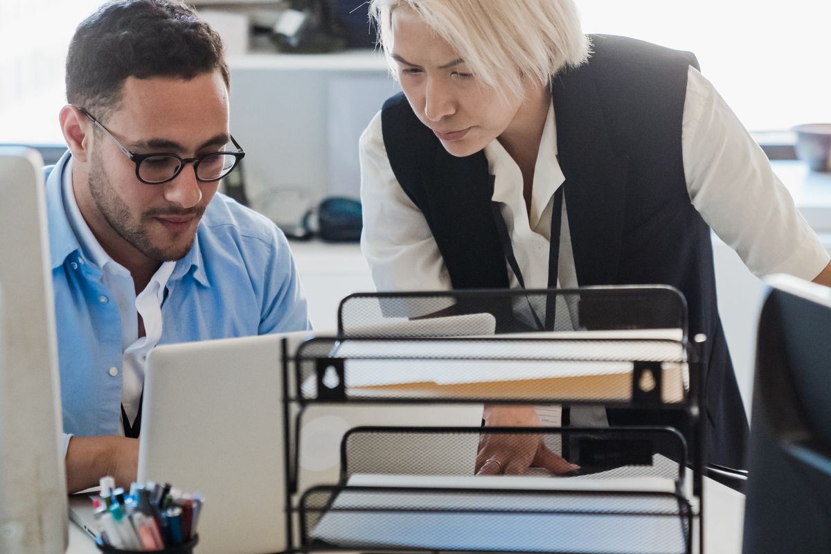 Two colleagues at a desk, one typing on a laptop while the other looks at the screen