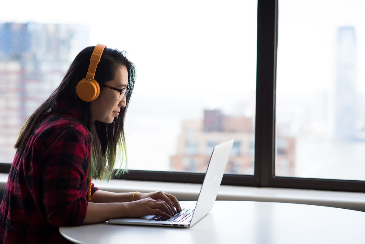 A woman wearing orange headphones works at a laptop beside a large city window