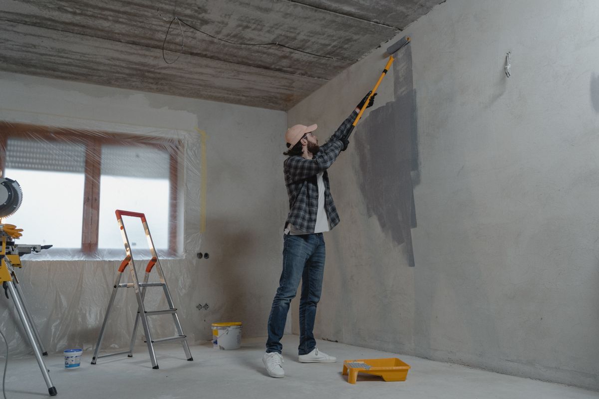 A decorator on a stepladder rolling grey paint onto a bare plaster wall in an empty room