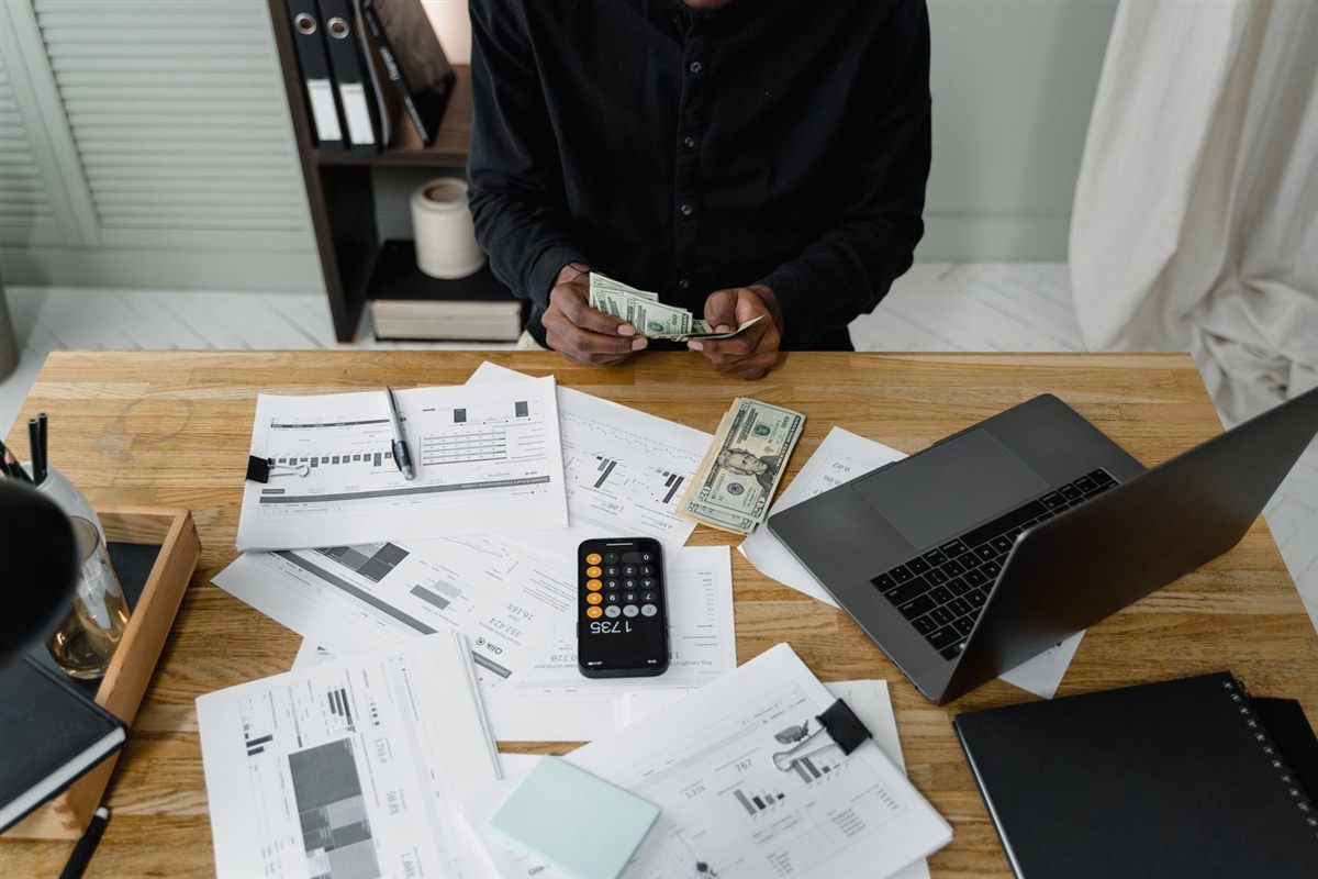 A business owner counts cash at a desk covered in reports and a laptop