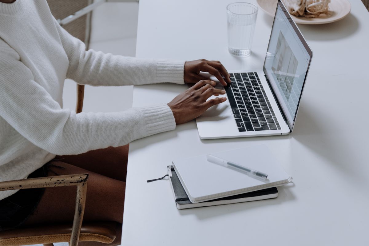 A person in a white jumper typing on a laptop at a white desk with a notebook and a glass of water