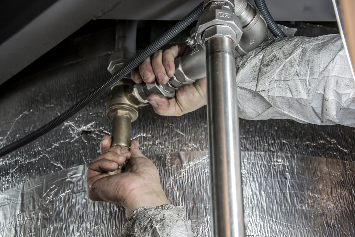A plumber tightening a brass pipe fitting by hand under a unit