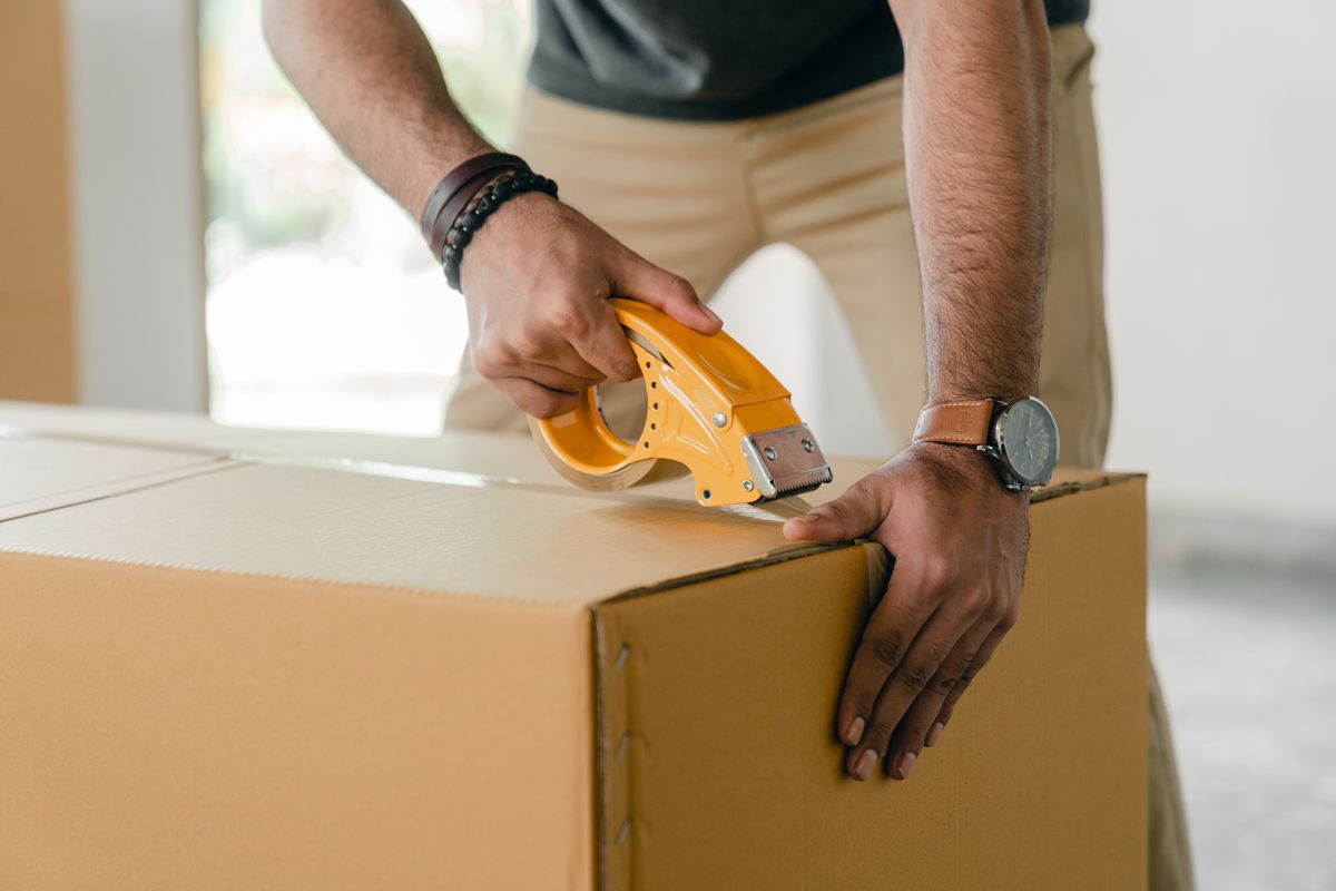 A man seals a large cardboard return box with a yellow tape dispenser