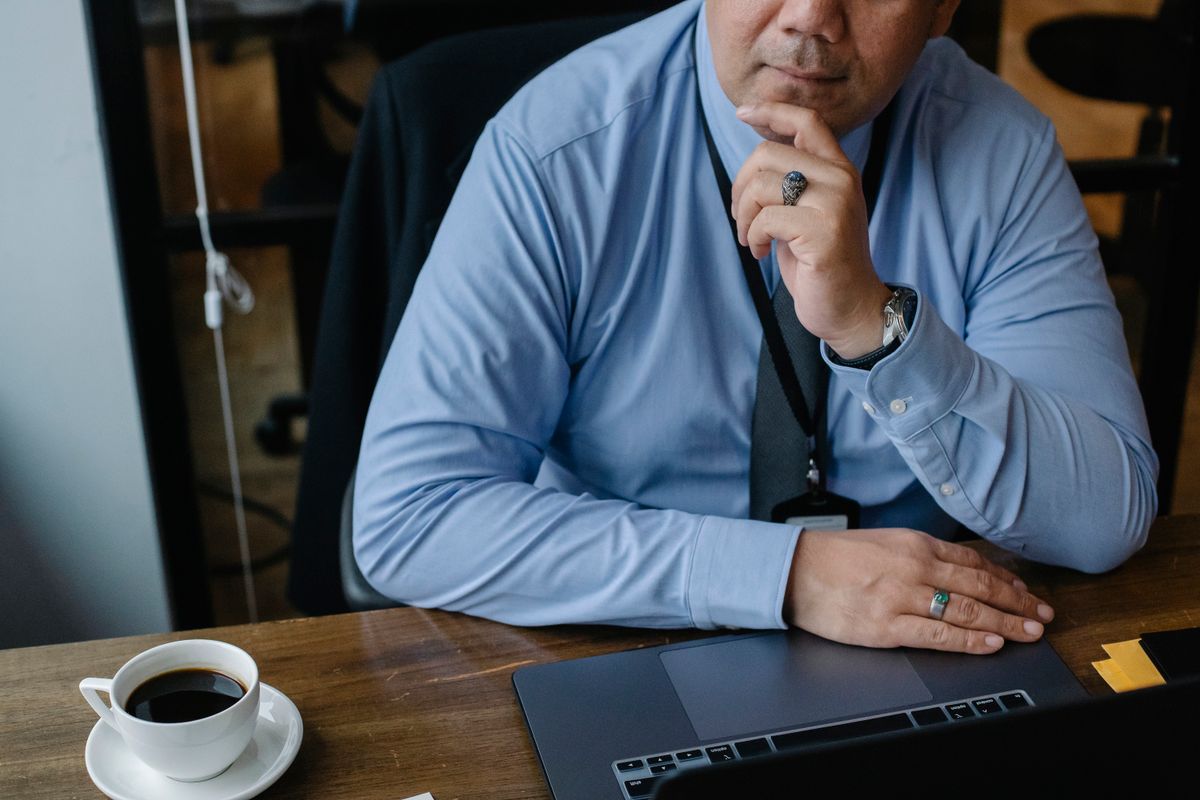 A business owner pauses with hand on chin before typing a reply at a laptop, a coffee on the desk