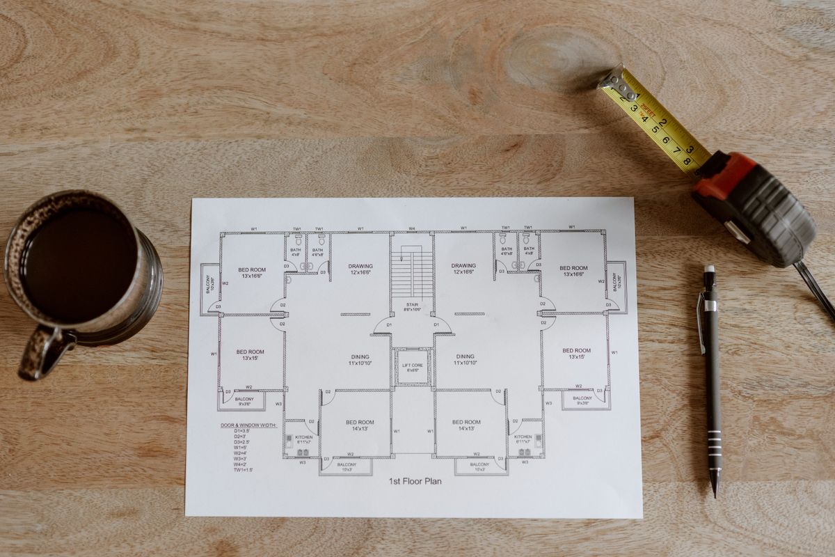 A floor plan on a wooden table beside a tape measure, a pencil and a cup of coffee