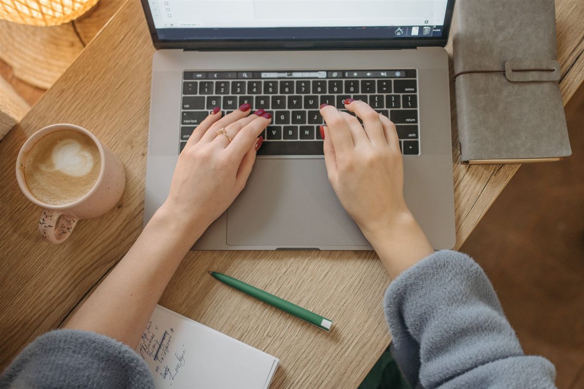 A person typing an email on a laptop beside a coffee and notebook