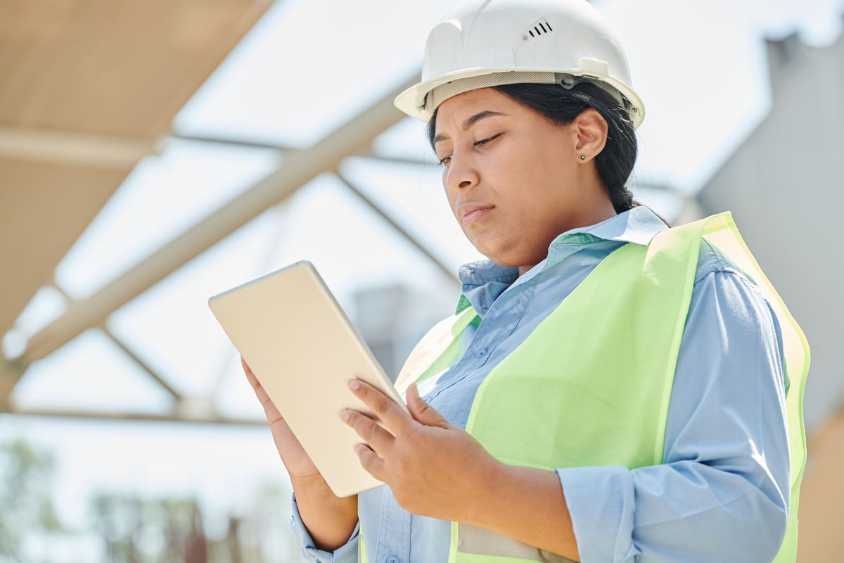 A construction worker in a white hard hat and hi-vis vest reading from a tablet on site