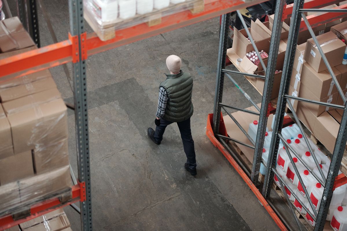 A worker walking a warehouse aisle between tall shelves of boxed stock