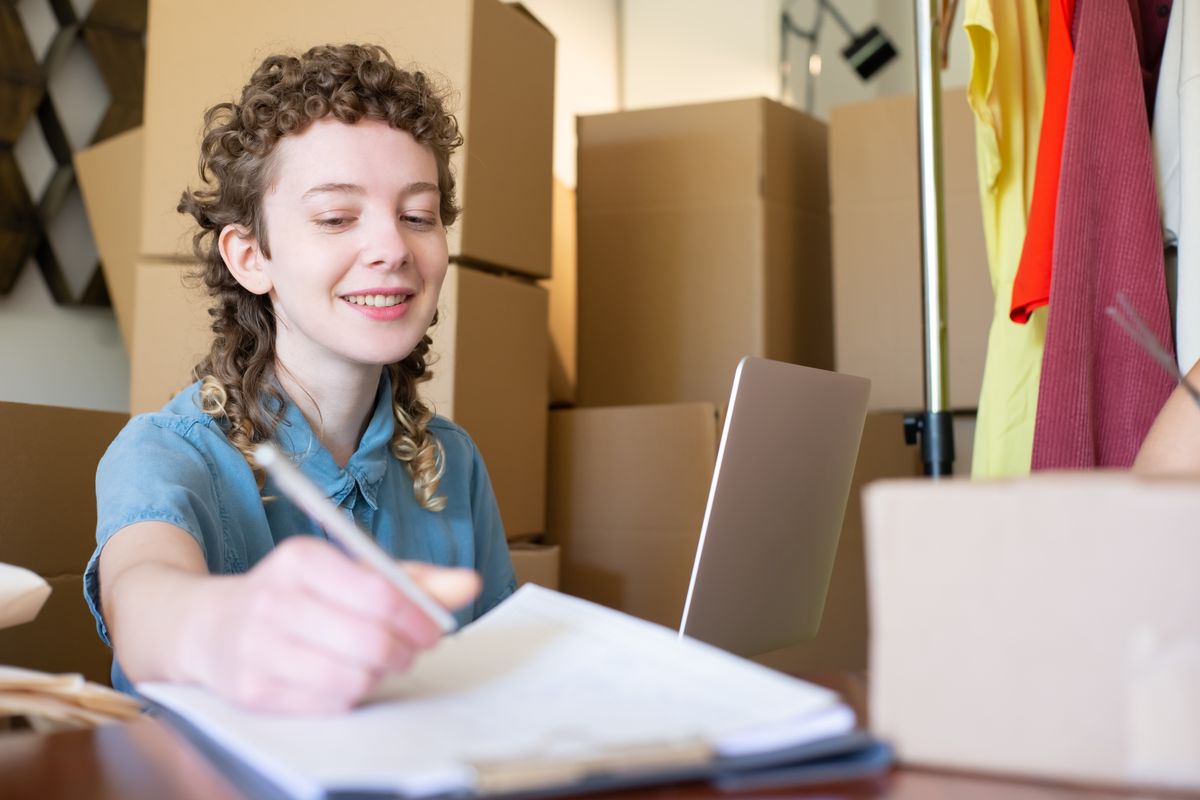 A store owner smiles while writing an order note at a desk surrounded by cardboard boxes and a laptop