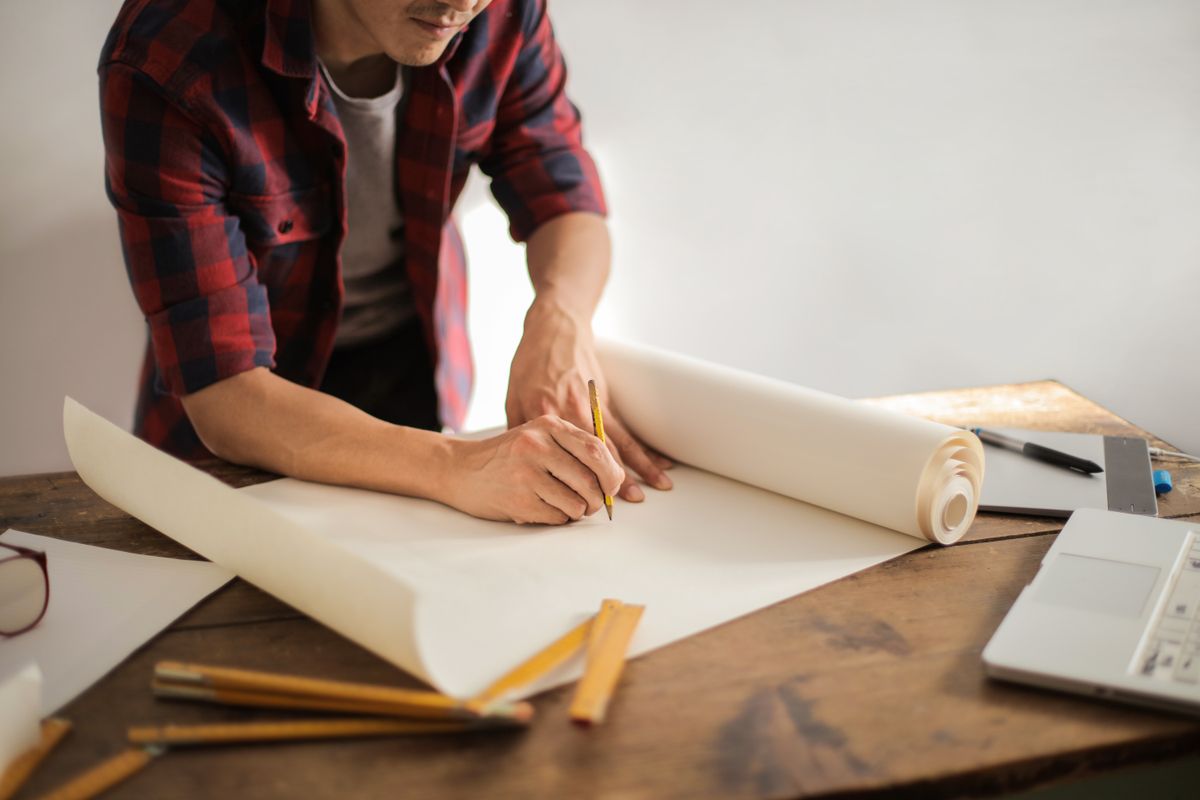 A tradesperson in a checked shirt sketching with a pencil on a roll of paper at a wooden workbench beside a laptop