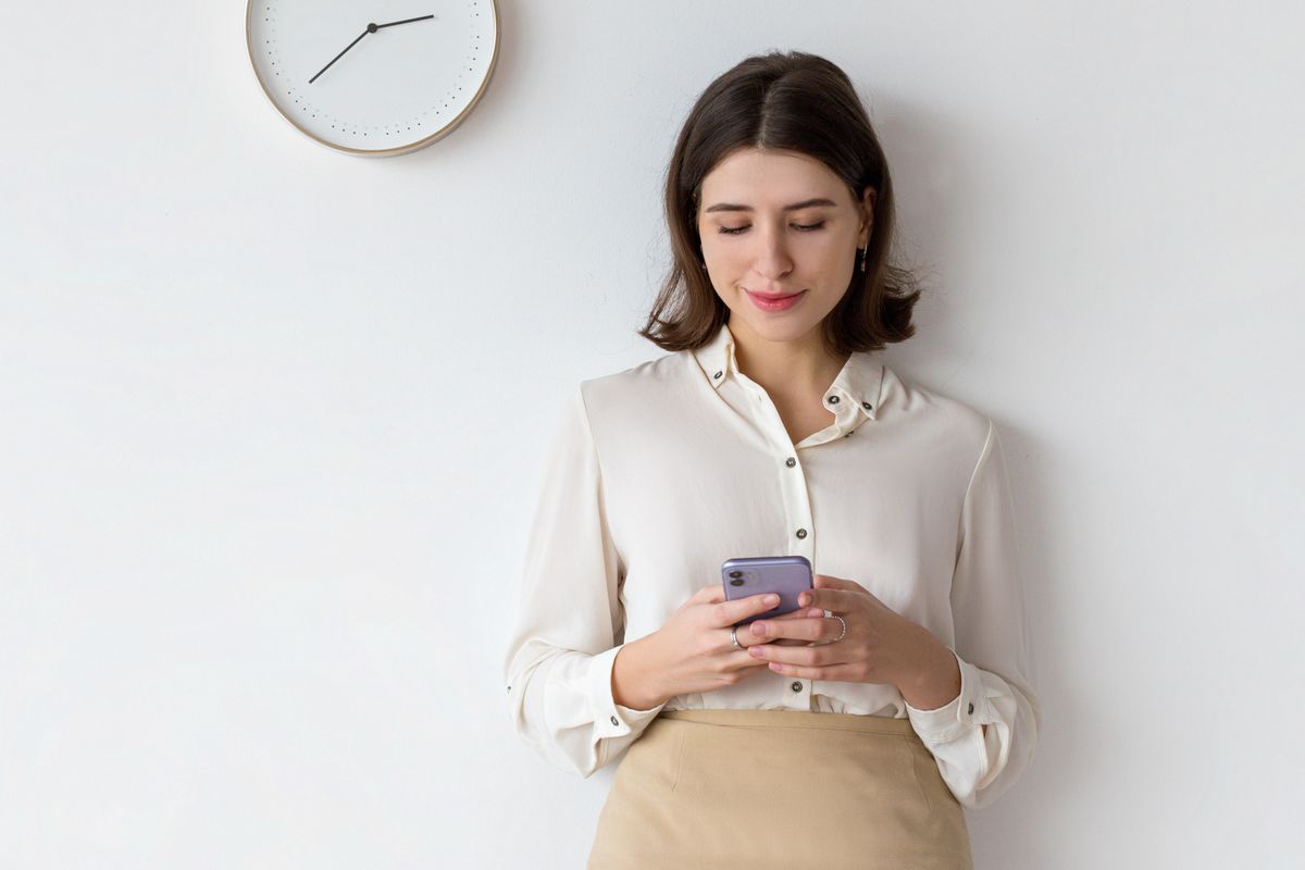 A woman checking her phone beneath a wall clock while she waits