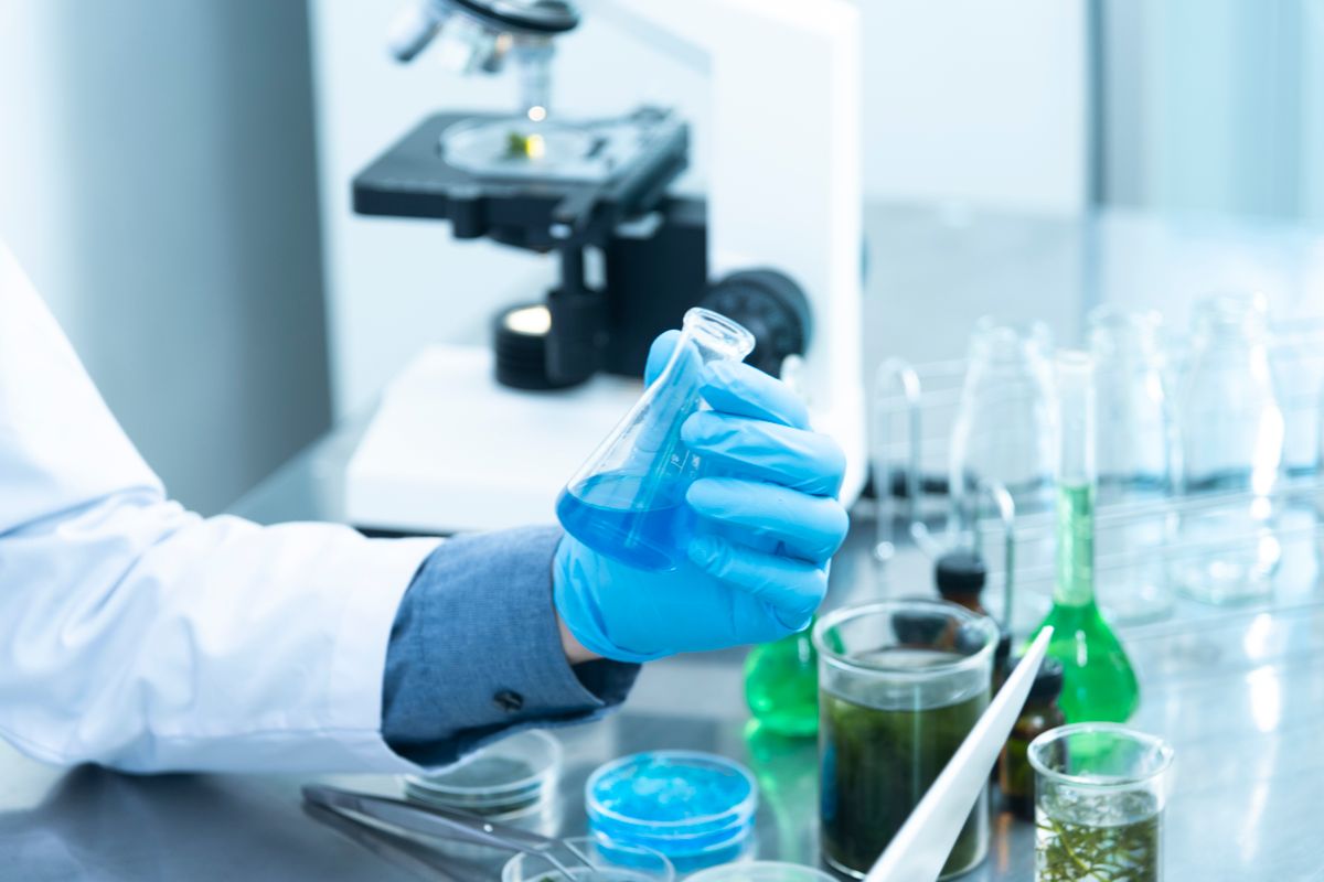 A gloved researcher holding a flask beside a microscope at a lab bench