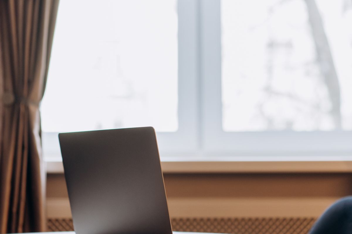 A laptop open on a table beside a bright window with a curtain drawn back