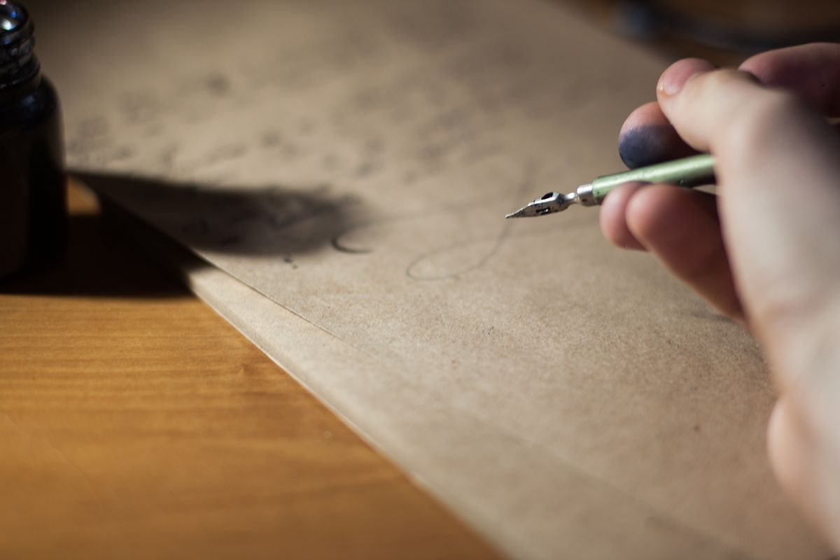 A hand writing with a dip pen on warm paper beside an ink bottle