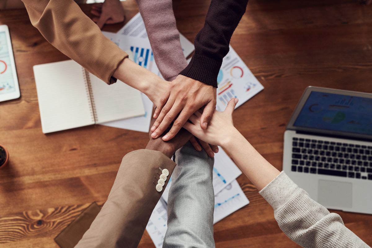 A small team joining hands over a desk covered in notes and a laptop